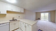 This single-floor residence offers a practical and functional layout with a clean, neutral aesthetic throughout. The kitchen is well-appointed with white cabinetry, light speckled countertops, a stainless steel sink, and tile flooring. A peninsula counter extends into the living area, creating an open-concept flow and additional workspace. White appliances—including a refrigerator, oven, and dishwasher—complete the kitchen's practical design. The living area is generously sized, featuring wall-to-wall carpeting, warm beige walls, and a textured ceiling. A sliding glass door leads to an outdoor balcony, allowing natural light to fill the space and providing an outdoor view. A wall-mounted HVAC unit ensures climate control convenience. The bedroom is spacious and carpeted, featuring a large window with vertical blinds that invites natural light and a wall-mounted HVAC unit. The room's open layout offers flexibility in furniture arrangement. The bathroom is finished with tile flooring and includes a full-size tub and shower combination enclosed by sliding glass doors with chrome hardware. Throughout the space, the consistent use of light, neutral tones creates a cohesive and bright atmosphere.