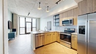 This thoughtfully designed residence offers four bedrooms and two bathrooms across a single floor, blending contemporary industrial aesthetics with warm, natural materials. The open-concept kitchen serves as the heart of the home, featuring light wood cabinetry, stainless steel appliances including a refrigerator with ice maker, electric range, built-in microwave, and dishwasher. A peninsula with a quartz countertop and seating provides a casual dining area, while open metal-framed shelving adds a modern industrial touch. The striking exposed concrete ceiling, pendant lighting, and track lighting further enhance the loft-inspired character of the space. Herringbone-patterned hardwood flooring flows throughout the main living areas, adding warmth and visual interest. Floor-to-ceiling windows flood the space with natural light and offer expansive city views. Each of the four bedrooms features sage green paneled accent walls, wood-platform beds with built-in storage drawers, and compact workstation areas. Select bedrooms enjoy direct access to city views through large windows. The two bathrooms are finished with wood-tone vanities, illuminated mirrors, light blue accent walls, walk-in glass-enclosed showers with rain and handheld fixtures, and textured stone-look tile flooring, delivering a clean and spa-inspired feel throughout.