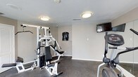 This single-level fitness center offers a functional and straightforward workout space designed to meet essential exercise needs. The room features neutral gray walls and dark rubberized flooring, providing a practical environment suited for physical activity. Two flush-mount ceiling light fixtures illuminate the space evenly, ensuring adequate visibility throughout.

The fitness center is equipped with a multi-function cable weight machine, which serves as the room's centerpiece, offering a variety of strength training exercises. A cardio machine, visible on the right side of the space, complements the strength equipment. A wall-mounted flat-screen television is positioned in the upper corner of the room, allowing users to stay entertained during workouts.

A posted fitness center rules sign is displayed on the wall, indicating that this is a shared or communal facility. The open floor area provides enough room for stretching or additional floor-based exercises. The space maintains a clean, no-frills aesthetic, prioritizing function over ornamentation, making it a straightforward and accessible option for those seeking a convenient fitness facility.