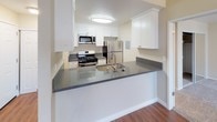 This single-story unit at 336 Pine Ave in Carlsbad offers a well-organized layout with a variety of functional living spaces. The floor plan, visible in the 3D model, reveals a thoughtfully arranged interior that balances open common areas with more private rooms.

The living area features rich hardwood-style flooring, a ceiling fan with integrated lighting, and a sliding glass door fitted with vertical blinds that opens to an outdoor patio, allowing natural light to fill the space. An adjacent room, carpeted for comfort, mirrors this setup with its own sliding glass door and ceiling fan, offering flexibility as a bedroom or secondary living space.

The kitchen stands out with its contemporary design, showcasing white shaker-style cabinetry, sleek gray countertops, a peninsula with an undermount sink, and a full suite of stainless steel appliances including a gas range, microwave, and refrigerator. Wood-look flooring runs throughout this area, complementing the clean aesthetic.

The bathroom features a full tub-shower combination with a clean white surround, chrome fixtures, and wood-style flooring. Built-in cabinetry provides practical storage. Throughout the unit, neutral wall tones and white trim create a cohesive, move-in-ready ambiance.