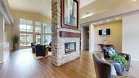 This single-story community clubhouse is designed to impress from the moment you enter. The centerpiece of the space is a dramatic floor-to-ceiling stacked-stone fireplace column that anchors the open great room, drawing the eye upward to soaring vaulted ceilings. Expansive floor-to-ceiling windows flood the lounge area with abundant natural light and provide views of the surrounding exterior, creating a bright and airy atmosphere throughout.

The lounge is comfortably appointed with plush leather seating, accent tables, and warm hardwood-style flooring that extends seamlessly throughout the space. A built-in bar area with counter seating and beverage service adds a social hub for residents, while a wall-mounted flat-screen television enhances the entertainment experience. Decorative potted plants and curated artwork contribute to the inviting, polished ambiance.

Tucked off the main lounge is a dedicated office area, thoughtfully designed with a large L-shaped desk, guest seating, and a bold teal accent wall adorned with framed artwork. Recessed lighting and a door with privacy blinds ensure a functional and professional environment. A console table with decorative accents near the entry adds a refined finishing touch. Overall, the space strikes a balance between comfort, style, and functionality.