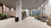 This single-floor space presents a thoughtfully designed lobby and lounge area that blends modern aesthetics with organic materials. The interior is anchored by a dramatic herringbone-patterned wood accent wall in warm, multi-toned hues, which serves as a striking visual focal point. Opposite this, floor-to-ceiling black-framed glass walls and doors flood the space with natural light while offering views of a lush planted corridor, seamlessly connecting the interior to its surroundings.

At the heart of the room sits a rectangular brick-clad water feature with a dark stone surround, adding a calming, architectural element to the open floor plan. The polished concrete flooring runs throughout, complementing the neutral gray tones of the walls and structural columns.

Seating is arranged in distinct zones, with rattan egg chairs, woven bistro chairs, and wooden café tables distributed throughout, offering both intimate nooks and casual gathering spaces. A dedicated dining area with a long table and rattan chairs occupies one corner, suitable for small group meetings or communal use.

Recessed ceiling lighting provides even, warm illumination across the entire space, while curated greenery — including large fiddle-leaf fig trees and tropical plantings — enhances the biophilic character of the environment. Overall, the space exudes a refined yet welcoming ambiance.