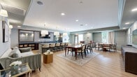 Located at 2545 Jessamine Grove Drive in Indian Land, South Carolina, this single-floor space is a well-appointed combination of dining and kitchen areas designed for both functionality and refined aesthetics. Throughout the space, warm light wood flooring provides a cohesive foundation, complemented by a soft sage-green color palette and detailed crown molding that lends an air of sophistication.

The dining area features elegant wainscoting panels along the walls, recessed lighting, and a large rectangular wood dining table surrounded by upholstered chairs — ideal for formal gatherings. A wall-mounted flat-screen television and a dark wood sideboard add both practicality and style. A separate card table area with a tufted leather accent chair provides an additional casual seating option, with a window offering views of an outdoor deck and greenery beyond.

The kitchen is a standout feature, showcasing a generously sized central island with a grey quartz countertop and seating for multiple guests along one side. Oversized pendant lights with brass fixtures hang above, creating a warm, inviting glow. The back kitchen area is highlighted by a striking geometric patterned tile backsplash, rich dark wood cabinetry, stainless steel sinks, and commercial-grade refrigeration, underscoring the space's capacity for hosting and entertaining.