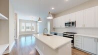 This well-appointed single-floor residence showcases a contemporary design with a cohesive neutral palette and modern gold hardware accents throughout. The heart of the home is an open-concept kitchen that flows seamlessly into the living area, anchored by a generous center island with a white quartz countertop, an undermount sink, and a sleek gold faucet. The kitchen is equipped with stainless steel appliances, including a gas range, over-the-range microwave, and a French door refrigerator. Upper white shaker cabinets are complemented by soft blue-gray lower cabinets and a chevron-patterned tile backsplash. Stylish cone-shaped pendant lights hang above the island, adding a designer touch. The living area features warm hardwood flooring, large windows that invite natural light, and a modern brass chandelier. A dedicated mudroom with built-in cubbies and hooks offers practical entryway storage. Bedrooms are carpeted for comfort and include ample natural light from well-positioned windows. The bathrooms are finished with large-format tile flooring, quartz vanity tops, gold fixtures, and matte black accents. The primary bathroom features a walk-in glass-enclosed shower with white subway tile walls and hexagonal mosaic tile flooring, accompanied by access to a spacious walk-in closet with wire shelving.