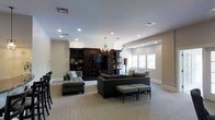 This single-floor space showcases a spacious, open-plan living area designed with a refined, contemporary aesthetic. The room is anchored by a substantial dark wood built-in wall unit, which houses a large flat-screen television and open shelving — serving as both a functional and visual centerpiece. Seating is arranged thoughtfully throughout, with multiple dark upholstered sofas and tufted ottomans creating distinct yet connected conversation areas across the patterned, light-toned carpeting.

Lighting is layered effectively, combining recessed ceiling fixtures that provide even ambient illumination with an elegant chandelier suspended at the room's center, adding a warm, decorative touch. The neutral beige walls and pale carpet keep the overall palette cohesive, while the darker furnishings introduce contrast and depth.

Architectural detailing includes arched doorway transitions and a subtly stepped ceiling design that defines different zones within the open layout. Natural light enters through multiple windows along one wall, fitted with white roller blinds. A dining area with dark chairs is partially visible, suggesting additional functional space within this well-appointed, single-level interior.