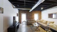 This single-floor loft space showcases a compelling industrial aesthetic that blends raw architectural elements with comfortable living. The exposed brick walls serve as a defining feature, adding warmth and texture to the space while lending an authentic urban character. The open ceiling reveals structural beams, black-painted joists, and prominent silver HVAC ductwork, creating a bold industrial overhead statement that adds considerable vertical depth to the interior.

Polished concrete floors run throughout, providing a sleek, durable foundation that complements the rugged charm of the brick and ceiling details. The living area is thoughtfully arranged with neutral-toned sofas, a rustic wooden coffee table, and a patterned area rug that softens the industrial elements underfoot. A media console with a flat-screen television anchors one end of the room.

Natural light fills the space generously through multiple tall, white-framed windows and a glass-paneled exterior door set directly into the brick wall, brightening the interior and highlighting the rich texture of the masonry. Track lighting mounted to the ceiling provides additional illumination. The overall ambiance strikes a balance between the rugged and the refined, making this loft a distinctive and characterful space.