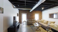 This single-floor loft space showcases a compelling industrial aesthetic that blends raw architectural elements with comfortable living. The exposed brick walls serve as a defining feature, adding warmth and texture to the space while lending an authentic urban character. The open ceiling reveals structural beams, black-painted joists, and prominent silver HVAC ductwork, creating a bold industrial overhead statement that adds considerable vertical depth to the interior.

Polished concrete floors run throughout, providing a sleek, durable foundation that complements the rugged charm of the brick and ceiling details. The living area is thoughtfully arranged with neutral-toned sofas, a rustic wooden coffee table, and a patterned area rug that softens the industrial elements underfoot. A media console with a flat-screen television anchors one end of the room.

Natural light fills the space generously through multiple tall, white-framed windows and a glass-paneled exterior door set directly into the brick wall, brightening the interior and highlighting the rich texture of the masonry. Track lighting mounted to the ceiling provides additional illumination. The overall ambiance strikes a balance between the rugged and the refined, making this loft a distinctive and characterful space.
