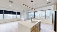 Located on the sixth floor at 411 N Akard St in the heart of Dallas, this 2-bedroom, 2-bathroom residence offers a refined urban living experience with high-end finishes throughout. The open-concept layout seamlessly connects the dining room, kitchen, and living room into one expansive, light-filled space, anchored by floor-to-ceiling windows that frame dramatic downtown Dallas skyline views. Light hardwood flooring runs throughout, complementing the crisp white walls and clean modern aesthetic.

The kitchen is a standout feature, centered on a generous marble-look quartz waterfall island with an integrated sink and brass fixtures. Taupe cabinetry with gold hardware pairs with stainless steel appliances, including a gas range and French door refrigerator. A linear pendant light adds a sophisticated focal point above the island.

Both bedrooms are well-proportioned and filled with natural light from large windows overlooking the city. The primary bedroom includes ample space and access to its own bathroom. Both bathrooms are finished with dramatic marble-style tile on floors and walls, warm taupe vanities, and gleaming gold hardware and fixtures, including a glass-enclosed shower in the primary bath and a soaking tub in the second.

Additional spaces include a hallway, laundry room, utility room, pantry, and two closets, ensuring excellent functionality throughout.