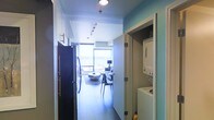 This single-floor space offers a thoughtfully designed open layout that maximizes functionality and flow. The entryway leads directly into a galley-style kitchen outfitted with stainless steel appliances, including a full-size refrigerator, set against clean white cabinetry. Track lighting above illuminates the corridor, creating a well-lit transition through the space. A dedicated in-unit laundry closet is conveniently positioned off the hallway, housing a stacked washer and dryer unit — a practical feature that adds everyday convenience. The hallway opens into a bright and airy living area, where large floor-to-ceiling windows flood the space with natural light and frame a stunning water view beyond. The living area features hardwood-style flooring throughout, lending warmth and continuity to the interior. The overall ambiance is modern and understated, with a neutral color palette accented by soft teal walls that add a subtle touch of personality. The open sightlines from the entryway to the living area create a sense of depth and spaciousness, making this a well-appointed and efficiently designed single-level residence.