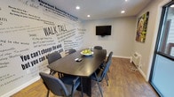 This single-floor meeting room is designed to inspire productivity and collaborative thinking. The space is anchored by a bold, typographic accent wall covered in motivational quotes and phrases — including prominent text such as "Walk Tall," "Think Outside the Box," and excerpts attributed to notable figures like Winston Churchill. The wall serves as both a design statement and a source of encouragement for occupants.

A dark, rectangular conference table sits at the center of the room, surrounded by cushioned task chairs that comfortably accommodate several attendees. A wall-mounted flat-screen television is positioned directly across from the seating arrangement, making it well-suited for presentations and collaborative screen sharing. Recessed ceiling lighting provides clean, even illumination throughout the space.

The flooring features warm, wood-look planks that add a grounded, professional feel, complementing the neutral gray tones of the walls and ceiling. The overall ambiance is energetic yet focused — a space intentionally curated to motivate and engage those who use it. Whether for team meetings, brainstorming sessions, or one-on-one discussions, this room offers a purposeful and visually stimulating environment for productive work.