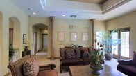 Villa Toscana is a single-floor space that exudes the warmth and sophistication of Tuscan-inspired design. The reception area serves as the centerpiece of the space, immediately drawing attention with its stately decorative columns finished in a rich, aged stone texture. The walls are adorned in soft sage green tones, lending a calm and inviting ambiance throughout. Prominently displayed signage for Villa Toscana and its sub-sections, including Villa Florentine, provides clear wayfinding while adding to the space's refined character. Recessed lighting in a coffered ceiling highlights the architectural detailing above, while natural light filters in through a nearby window, brightening the space and adding warmth. The reception lounge features richly upholstered seating with ornate scrollwork detailing, complemented by fresh floral arrangements and decorative potted plants that bring life and color to the interior. Framed artwork lines the feature wall, contributing to the gallery-like atmosphere. A wide arched corridor leads further into the facility, hinting at additional spaces beyond. Overall, Villa Toscana presents a polished, welcoming environment with thoughtful design details throughout.