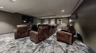 Located at 1600 Harbor Blvd in Weehawken, NJ, this dedicated screening room delivers an upscale cinematic experience within a thoughtfully designed, single-level space. The room is furnished with rows of generously sized brown leather recliner chairs, each equipped with built-in cup holders, arranged in a theater-style configuration to ensure optimal sightlines for every seat. Two flat-screen displays are mounted on opposing walls, offering flexible viewing options throughout the room. The walls are finished in a sophisticated gray textured treatment, complemented by soft, recessed ceiling lighting and wall sconces that create an intimate, cinema-like ambiance. A decorative built-in credenza with frosted glass cabinet doors adds a refined storage element along one wall, while a series of framed artwork pieces contributes to the polished aesthetic. The flooring features a bold, geometric-patterned carpet in shades of gray, black, and gold, adding visual depth and warmth to the space. Subtle details such as a decorative wall medallion and carefully placed accent lighting further enhance the room's character. The overall design balances comfort and style, making this screening room a well-appointed venue for private viewings and entertainment gatherings.