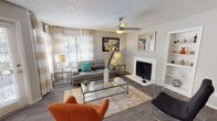 This single-floor space centers around a well-appointed living area that balances comfort and contemporary style. The room features hardwood-style flooring throughout, topped with a textured area rug that anchors the seating arrangement. A sleek gray sofa with teal accent pillows faces a glass-top coffee table with a metallic frame, while vibrant orange armchairs introduce a bold pop of color to the neutral palette.

A white fireplace serves as a focal point along one wall, flanked by wall art that adds visual interest to the space. Opposite, large windows dressed with sheer white curtains and horizontal blinds allow natural light to fill the room, creating a bright and airy atmosphere. A ceiling fan with an integrated light fixture provides both ventilation and additional illumination.

The walls are painted in a soft gray tone, complementing the textured ceiling finish. Decorative elements, including a tall white floor vase with dried branches, glass-and-metal side tables, and framed artwork, contribute to a cohesive, modern aesthetic throughout the living space.