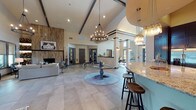 Located at 1747 FM 1101 in New Braunfels, Texas, this single-floor community clubhouse offers an impressive open-concept layout defined by soaring vaulted ceilings with exposed dark wood beams and dramatic chandelier lighting. Large-format tile flooring flows throughout the space, anchoring the expansive main gathering area where a central pedestal table serves as a natural focal point beneath a statement wrought-iron chandelier.

The full kitchen is outfitted with dark espresso cabinetry, granite countertops, stainless steel appliances, and a white subway tile backsplash. A granite-topped bar counter with barstool seating extends into the common area, illuminated by cluster pendant lights. Multiple lounge areas provide varied seating arrangements, including a cozy fireside nook featuring a wood-panel accent wall, built-in shelving stacked with firewood, and a fireplace — creating a warm, rustic ambiance.

Additional seating zones offer tufted leather chairs and patterned area rugs, bathed in natural light through large windows dressed with woven roller shades and teal drapery panels. The entry vestibule features floor-to-ceiling glass double doors on both ends, flooding the corridor with daylight and views of the surrounding grounds. A popcorn station and bistro-style high-top table add a playful, community-oriented touch to this well-appointed gathering space.