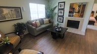 This single-floor space offers a warm and inviting atmosphere, highlighted by rich hardwood flooring that flows throughout the main living area. The living room serves as the focal point of the home, showcasing a tile-surround fireplace that adds both charm and comfort to the space. A large window allows abundant natural light to fill the room, creating a bright and airy feel. The layout is thoughtfully designed to accommodate comfortable seating and functional living, with ample room for furniture arrangement. Lush indoor plants enhance the space with a touch of greenery, complementing the neutral, earthy color palette of the walls and furnishings. Wall art displayed in a vertical gallery arrangement adds a decorative element and a sense of curated style. The transition between the hardwood flooring and the tile near the fireplace adds visual interest and clearly defines the living area. Overall, this space combines practical design with cozy, livable touches, making it a comfortable and functional single-level home.