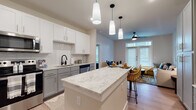 Located along Ed Bluestein Boulevard in Austin, TX, this single-floor residence offers a thoughtfully designed open-concept layout with contemporary finishes throughout. The kitchen is a standout feature, showcasing two-tone cabinetry with white upper cabinets and gray lower cabinets, a classic subway tile backsplash, granite countertops, and a generous central island with bar seating. Stainless steel appliances — including a refrigerator, range, microwave, and dishwasher — complete the fully equipped cooking space. Pendant lighting above the island adds a refined touch, while recessed lighting ensures the entire area is well lit. The kitchen flows seamlessly into the dining and living areas, which are anchored by light wood-style flooring and a ceiling fan. French doors in the living room invite natural light and provide access to the outdoors. The bedroom is carpeted and comfortably proportioned, featuring multiple windows dressed with blinds and dark curtains, a ceiling fan, and ample wall space. The en-suite bathroom is appointed with gray cabinetry, granite countertops, a dual-sink vanity with matte black fixtures, a large mirror, and tile flooring, directly accessible from the bedroom for added convenience.