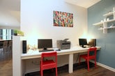 This single-floor business center is thoughtfully designed to offer a functional and visually appealing workspace. The layout features a welcoming entryway corridor with smooth white walls, vaulted ceiling accents, and rich dark hardwood flooring that extends throughout the space. Warm wall-mounted sconces and task lighting create an inviting ambiance as you move through the space.

The main work area is anchored by a long white quartz-style countertop workstation running along the wall, accommodating multiple computer terminals, keyboards, and a Brother multifunction printer, making it well-equipped for everyday business tasks. Vibrant red chairs add a bold pop of color against the neutral tones of the room. A colorful abstract painting mounted above the workstation serves as a standout decorative element, infusing the space with creative energy.

A secondary alcove workspace features a teal accent wall adorned with white floating shelves displaying tasteful décor pieces, alongside an additional computer workstation and desk. Natural light filters in through windows visible in the background, connecting the interior to the greenery outside. A reception-style counter near the entry provides informational materials and brochures, rounding out this well-appointed and efficiently organized business center.