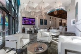 Located in Atlanta's prestigious 30326 corridor, this single-floor commercial space makes a striking first impression with its dramatic double-height ceilings and expansive floor-to-ceiling glass facade that floods the interior with abundant natural light and provides open views of the surrounding greenery. The architecture balances clean white wall panels with warm wood accents, creating a refined yet welcoming atmosphere throughout.

The main lounge area features a mix of glossy white tile flooring and distinctive pebble-mosaic carpeting, anchored by sleek white upholstered seating arranged around chrome-accented tables. Gold geometric pendant chandeliers hang in clusters overhead, adding an elegant warmth to the bright, airy environment. Large-scale contemporary artwork punctuates the white walls, contributing to the space's gallery-like sensibility.

A dedicated workstation area showcases a bold teal accent wall adorned with sculptural metallic panels. A slatted wood ceiling with recessed track lighting and a cascading chandelier distinguish this zone, which features a long reception-style desk with multiple computer workstations. Chrome railings define level transitions, while additional seating areas offer comfortable spots throughout, making this space as functional as it is visually compelling.