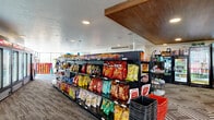 This single-floor convenience store offers a generously sized retail environment thoughtfully designed for both functionality and customer comfort. The main retail area features rows of well-stocked black merchandise shelving units displaying a wide variety of snacks, chips, and packaged goods, making product browsing easy and intuitive. A distinctive wood-paneled drop ceiling accent adds a warm architectural touch, contrasting with the textured white ceiling throughout the rest of the space. Underfoot, durable patterned carpet tiles in neutral tones provide a clean, commercial aesthetic, with a bold geometric-design area rug anchoring one section of the floor. Along the perimeter, a robust lineup of glass-door refrigeration units keeps cold beverages and perishables accessible and well-organized. Recessed lighting throughout ensures bright, even illumination across the entire retail floor. A separate secondary area transitions into a lounge-style space featuring floor-to-ceiling windows that flood the room with natural light and offer open exterior views. This area includes casual seating, a beverage fountain station, additional refrigeration, and a large-format Connect Four game, creating an inviting, relaxed atmosphere. Together, the two areas form a versatile and welcoming retail destination.