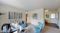 This single-floor residence offers a comfortable and thoughtfully arranged living environment with a clean, modern aesthetic. The living area serves as the heart of the home, featuring plush wall-to-wall carpeting and an abundance of natural light that fills the space with warmth throughout the day. Soft, neutral walls provide a versatile backdrop, complemented by contemporary artwork that adds subtle pops of color and personality. The room comfortably accommodates a seating arrangement centered around a stylish coffee table, creating an inviting atmosphere for relaxation and entertaining. A partial dividing wall adds architectural interest while maintaining an open, airy feel as the living space flows naturally toward the dining area, which is visible in the background and features hard flooring for a pleasing contrast in texture. Additional rooms are accessible via a hallway, suggesting a well-organized layout designed for everyday functionality. Ceiling-mounted lighting ensures the space remains well-lit, and clean baseboards and trim throughout contribute to the home's polished, well-maintained appearance. Overall, the residence exudes a bright, welcoming ambiance that balances practicality with a contemporary sense of style.