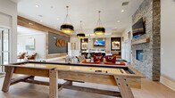 The Reserve at Kirkwood's clubroom is a thoughtfully designed community gathering space that seamlessly blends modern aesthetics with warm, inviting elements across a single open floor plan. The main lounge area anchors the space with bold, statement pendant lighting — featuring striking black-and-gold drum fixtures — suspended over a central wet bar outfitted with a white quartz countertop, bar seating, and a wall-mounted flat-screen TV framed in warm wood paneling. Equestrian-themed artwork adds a cohesive decorative thread throughout. A comfortable seating arrangement of plush sofas and vibrant rust-red accent chairs creates an inviting social hub, complemented by a rustic stone fireplace along one wall. A full-size shuffleboard table serves as the centerpiece of the game area, providing a focal point for recreation. Large glass doors flood the space with natural light and provide direct access to an outdoor patio. A high-gloss, rich red tile accent wall adds dramatic contrast near a secondary bar-height counter with modern white seating. A private conference room, enclosed with glass panels, offers an intimate setting with leather swivel chairs, a round table, textured carpet, and a wall-mounted display — ideal for small meetings or focused work sessions. Neutral large-format tile flooring, shiplap wall detailing, and curated artwork complete this polished yet comfortable community space.