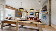 The Reserve at Kirkwood's clubroom is a thoughtfully designed community gathering space that seamlessly blends modern aesthetics with warm, inviting elements across a single open floor plan. The main lounge area anchors the space with bold, statement pendant lighting — featuring striking black-and-gold drum fixtures — suspended over a central wet bar outfitted with a white quartz countertop, bar seating, and a wall-mounted flat-screen TV framed in warm wood paneling. Equestrian-themed artwork adds a cohesive decorative thread throughout. A comfortable seating arrangement of plush sofas and vibrant rust-red accent chairs creates an inviting social hub, complemented by a rustic stone fireplace along one wall. A full-size shuffleboard table serves as the centerpiece of the game area, providing a focal point for recreation. Large glass doors flood the space with natural light and provide direct access to an outdoor patio. A high-gloss, rich red tile accent wall adds dramatic contrast near a secondary bar-height counter with modern white seating. A private conference room, enclosed with glass panels, offers an intimate setting with leather swivel chairs, a round table, textured carpet, and a wall-mounted display — ideal for small meetings or focused work sessions. Neutral large-format tile flooring, shiplap wall detailing, and curated artwork complete this polished yet comfortable community space.