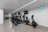 Located at 77 Parkwoods Village Dr in Toronto's Parkwoods-Donalda neighbourhood, this fitness centre offers a thoughtfully designed workout space across a single floor. The interior features a clean, modern aesthetic with crisp white walls, a speckled neutral-toned floor, and sleek linear LED lighting that runs along the ceiling to provide bright, even illumination throughout. A horizontal strip window along one wall brings in a touch of natural light, adding to the airy atmosphere.

The space is well-stocked with a diverse selection of exercise equipment. Cardio offerings include multiple treadmills and elliptical trainers, each equipped with individual monitors for entertainment during workouts. A recumbent bike provides an additional low-impact option. For strength training, a multi-function cable machine is available alongside a dumbbell rack and adjustable benches. Stability and flexibility training is supported by a dedicated ball rack stocked with various sizes of exercise balls.

Practical amenities include a wall-mounted water fountain with a bottle-filling station, wall-mounted televisions, and a wall-mounted air conditioning unit for climate control. A bold typographic motivational mural anchors one end of the room, reinforcing the energetic atmosphere. The space is rounded out by clearly marked emergency exits, ensuring a safe and functional environment for all users.