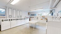 Located at 494 Avenue Rd in Toronto, this shared laundry room is a well-organized, functional facility designed to serve building residents efficiently. The space features a clean, bright interior finished in white throughout, with durable terrazzo flooring that is both practical and easy to maintain. Fluorescent lighting runs along the ceiling, ensuring the room is well-lit at all times.

Along one wall, a row of front-loading washing machines is neatly aligned, complemented by a separate bank of top-loading dryers paired with white overhead storage cabinets. A central folding table provides ample workspace for residents to sort and fold laundry. A utility sink with cabinetry is conveniently positioned along one wall, adding practical functionality to the space.

Windows along the perimeter allow natural light to filter in, contributing to the room's open and airy atmosphere. A community bulletin board keeps residents informed with notices and building updates. A charming bookshelf stocked with a wide variety of books adds a welcoming, community-oriented touch, giving residents something to enjoy while they wait. Additional amenities include a wall clock, fire safety equipment, and clearly marked entry and exit doors, making this laundry room a thoughtfully equipped and resident-friendly shared facility.