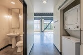This single-floor loft showcases a bold industrial aesthetic defined by exposed black steel structural beams, corrugated metal ceilings, and polished concrete floors throughout. The expansive main living area is the centerpiece of the space, bathed in abundant natural light streaming through large floor-to-ceiling windows that offer treetop views of the surrounding urban landscape. The open-plan layout provides a versatile, airy environment with generous proportions and a clean, minimalist feel.

The kitchen is thoughtfully designed with dark cabinetry contrasted against stainless steel appliances, including a range, microwave, dishwasher, and refrigerator. Dark countertops and a stainless steel sink complete the sleek, functional workspace, which flows seamlessly into the main living area via polished concrete flooring.

The bathroom features a classic pedestal sink, toilet, and a large mirror framed by industrial-style cage pendant lighting, maintaining the space's cohesive aesthetic. A dedicated laundry closet with a stacked washer and dryer unit adds practical convenience. A corridor connects the key areas of the space, with a barn-style sliding door providing privacy where needed. The combination of raw materials, structural transparency, and generous natural light creates a striking, contemporary atmosphere throughout.