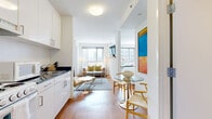Located on the 19th floor of 1188 Mission Street in San Francisco's SoMa neighborhood, this modern unit offers a thoughtfully designed open-concept layout bathed in natural light. Warm hardwood flooring flows throughout the main living areas, complementing the crisp white walls and clean contemporary aesthetic.

The kitchen is outfitted with white cabinetry extending to the ceiling, dark countertops, and a full suite of appliances including a refrigerator, electric range, microwave, and sink — offering ample functionality in a compact, efficient footprint. Adjacent to the kitchen, a glass-top dining table with woven wishbone chairs provides an intimate dining space, accented by a bold, colorful abstract artwork on the wall.

The living area features large floor-to-ceiling windows that frame dynamic city views of the San Francisco skyline, flooding the space with natural light. A wall-mounted flat-screen TV is positioned on a wood ledge between the living and dining areas, serving both zones effectively.

The bedroom is generously proportioned with a full-size bed and built-in closet storage. The bathroom is finished with white tile, a soaking tub with shower, built-in shelving for storage, a dark-countertop vanity, and a large mirror, maintaining the unit's consistent clean and modern design language throughout.
