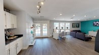 This single-floor space offers a clean, contemporary design that balances functionality with a welcoming ambiance. The open layout is anchored by smooth hardwood-style flooring in a warm gray tone, which runs seamlessly throughout the main area, contributing to a cohesive and polished look.

Natural light is a defining feature of this space, pouring in through a set of classic French doors and multiple large windows along the perimeter. These glazed openings frame outdoor views and create an airy, well-lit environment throughout the day. The white-painted walls and light ceiling amplify the brightness, giving the interior a fresh and spacious feel.

A dedicated workstation area with a white desk and seating provides a practical workspace, while a comfortable lounge area with a gray sofa offers a space for relaxation or informal gatherings. A white open-shelving unit adds both storage and decorative interest near the entrance, displaying tasteful accents.

Overhead, a combination of recessed lighting and an adjustable track lighting fixture ensures versatile illumination options to suit different needs and times of day. The overall design is modern yet approachable, making this a highly functional space suitable for a variety of professional or community uses.