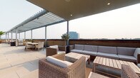 Located at 797 N Wall St in the heart of Columbus, Ohio, this space offers a compelling blend of indoor and outdoor environments. The standout feature is a generously sized rooftop terrace that delivers sweeping views of the Columbus skyline. The outdoor area is thoughtfully designed with a combination of covered and open sections, anchored by a modern pergola structure that provides shade while maintaining an open, airy atmosphere. The terrace is furnished with wicker lounge chairs and a full outdoor sofa set with neutral gray cushions, arranged around a wooden coffee table to create an inviting seating area. A separate dining arrangement is positioned nearby, offering additional space for gathering. A potted olive tree adds a touch of greenery and a Mediterranean aesthetic to the rooftop setting. The interior portions of the space feature windows with horizontal blinds, allowing for controlled natural light. The overall ambiance of the space is modern and urban, with clean lines and a neutral palette that complements the surrounding cityscape. This property is well-suited for those seeking a contemporary setting with exceptional outdoor amenities in a prime downtown Columbus location.