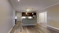 This single-story space showcases an open-concept layout that seamlessly connects the kitchen and living areas, creating a spacious and cohesive environment. The kitchen serves as the focal point, featuring rich dark-stained cabinetry that contrasts elegantly with a light-colored granite or stone countertop. A central island with a gray base provides additional workspace and acts as a natural dividing point between the kitchen and the adjoining living area. Stainless steel appliances, including a refrigerator, microwave, and range, contribute a polished, contemporary feel. Pendant lights hang above the island, offering targeted task lighting that enhances the kitchen's modern aesthetic, while recessed ceiling lights illuminate the broader space. Crown molding runs along the ceiling perimeter, adding a refined architectural detail. Warm-toned, hardwood-style flooring extends throughout the entire space, tying the rooms together cohesively. Multiple doorways are visible, suggesting additional rooms beyond the main open area. The neutral gray wall tones and white trim create a clean, versatile backdrop that complements the overall contemporary design.