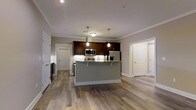 This single-story space showcases an open-concept layout that seamlessly connects the kitchen and living areas, creating a spacious and cohesive environment. The kitchen serves as the focal point, featuring rich dark-stained cabinetry that contrasts elegantly with a light-colored granite or stone countertop. A central island with a gray base provides additional workspace and acts as a natural dividing point between the kitchen and the adjoining living area. Stainless steel appliances, including a refrigerator, microwave, and range, contribute a polished, contemporary feel. Pendant lights hang above the island, offering targeted task lighting that enhances the kitchen's modern aesthetic, while recessed ceiling lights illuminate the broader space. Crown molding runs along the ceiling perimeter, adding a refined architectural detail. Warm-toned, hardwood-style flooring extends throughout the entire space, tying the rooms together cohesively. Multiple doorways are visible, suggesting additional rooms beyond the main open area. The neutral gray wall tones and white trim create a clean, versatile backdrop that complements the overall contemporary design.