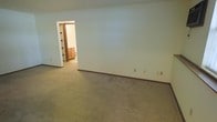This single-floor space presents a straightforward and functional layout designed for comfortable everyday living. The main living area is finished with wall-to-wall carpet in a neutral beige tone, lending the space a warm and understated feel. Smooth, light-painted walls are trimmed with simple baseboards that frame the room cleanly, contributing to a tidy and cohesive aesthetic throughout.

An open doorway connects the main living area to an adjoining room, through which warm wood cabinetry is visible, suggesting a kitchen or utility space with traditional wood-finished storage. The transition between rooms is seamless, offering an easy flow that makes the layout feel connected and accessible.

Electrical outlets are evenly distributed along the walls, providing practical convenience throughout the space. The overall interior palette is kept neutral, with soft tones across both the flooring and walls, creating a calm and move-in-ready environment. The absence of excess ornamentation gives the space a clean canvas quality, making it highly adaptable to a variety of furnishing styles and personal preferences. This single-level layout offers simplicity and ease of navigation, making it a practical and versatile space.