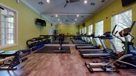 This fitness center is a thoughtfully designed, single-floor exercise facility finished in a vibrant sage green that lends the space an energizing atmosphere. The room features warm hardwood-style flooring throughout, crown molding along the ceiling, and recessed lighting complemented by ceiling fans that keep the environment comfortable during workouts.

A full-length mirrored wall runs along one side of the room, visually expanding the space and providing exercisers with proper form feedback. Multiple windows with horizontal blinds line the walls, allowing ample natural light to filter in while offering views of surrounding greenery.

The cardio section is well-stocked with a row of treadmills, elliptical trainers, and a stationary recumbent bike, all positioned for easy access. A dedicated strength training area includes a multi-station cable machine, a weight bench, and a full dumbbell rack with various weight increments, along with exercise mats and stability balls for flexibility and core work.

Wall-mounted televisions are positioned for viewing during cardio sessions. The facility also includes practical amenities such as a sanitizing dispenser, a water cooler with cup dispenser, a waste bin station, and a scale. A connecting hallway leads to additional areas of the broader facility, making this fitness center a convenient and fully equipped space for a comprehensive workout routine.