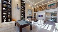 This upscale amenity space is defined by its dramatic double-height ceilings and refined contemporary design throughout. The central gathering area commands attention with a full-size billiards table positioned beneath a cascading cluster of globe pendant lights, creating a striking focal point. Herringbone-patterned wall panels in warm neutral tones line the walls, complemented by a sleek linear fireplace flanked by dark-stained open shelving units displaying curated décor. Adjacent lounge seating — including curved sofas and leather accent chairs — creates an inviting social atmosphere, while a wall-mounted TV and elevated mezzanine level overlook the space from above.

Large floor-to-ceiling windows and glass doors connect the billiards lounge directly to an outdoor pool area, flooding the interior with natural light. A separate lounge nook features intimate banquette-style seating with velvet chairs and artful wall sconces in a warm, bistro-inspired setting.

The full kitchen is outfitted with dark cabinetry, a marble-topped island with upholstered bar seating, stainless steel double ovens, and a bold stone backsplash beneath a stainless range hood. A bright dining area nearby features marble-topped tables and richly upholstered chairs beneath elegant pendant and chandelier lighting, rounding out this thoughtfully appointed communal environment.