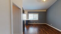 This single-floor residence showcases a cohesive and modern design aesthetic, highlighted by rich, dark hardwood flooring that runs throughout the entire space and ties each room together seamlessly. The entry area opens into a bright living space with slate-blue accent walls, crisp white trim, and soaring ceiling heights that lend an airy, open feel. A wall-mounted climate control unit provides practical comfort near the front entry.

The kitchen is a standout feature, outfitted with deep navy shaker-style cabinetry accented by gold hardware and glass-front upper cabinets. Light quartz countertops provide a striking contrast, while a full suite of stainless steel appliances — including a gas range, over-the-range microwave, dishwasher, and refrigerator — ensures a fully functional cooking space. A window above the sink allows natural light to fill the room.

The bedrooms continue the slate-blue color palette and are bathed in natural light through blinded windows. One bedroom features impressively tall ceilings and a built-in navy wardrobe with full mirrored sliding doors, adding both depth and storage. The hallway connecting the rooms is accented by a geometric pendant light fixture, adding a touch of contemporary style. The bathroom includes tile surround detailing and clean, neutral finishes throughout.