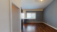 This single-floor residence showcases a cohesive and modern design aesthetic, highlighted by rich, dark hardwood flooring that runs throughout the entire space and ties each room together seamlessly. The entry area opens into a bright living space with slate-blue accent walls, crisp white trim, and soaring ceiling heights that lend an airy, open feel. A wall-mounted climate control unit provides practical comfort near the front entry.

The kitchen is a standout feature, outfitted with deep navy shaker-style cabinetry accented by gold hardware and glass-front upper cabinets. Light quartz countertops provide a striking contrast, while a full suite of stainless steel appliances — including a gas range, over-the-range microwave, dishwasher, and refrigerator — ensures a fully functional cooking space. A window above the sink allows natural light to fill the room.

The bedrooms continue the slate-blue color palette and are bathed in natural light through blinded windows. One bedroom features impressively tall ceilings and a built-in navy wardrobe with full mirrored sliding doors, adding both depth and storage. The hallway connecting the rooms is accented by a geometric pendant light fixture, adding a touch of contemporary style. The bathroom includes tile surround detailing and clean, neutral finishes throughout.