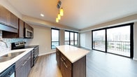 This contemporary single-floor space showcases a thoughtfully designed open-concept layout that seamlessly connects the kitchen and living areas. The kitchen is a standout feature, centered around a sleek island with a crisp white quartz countertop and dark cabinetry accented with gold hardware. Pendant lighting with warm-toned globes hangs above the island, adding an inviting ambiance. Stainless steel appliances, including a microwave and range, complete the modern culinary setup.

Wide-plank hardwood flooring flows throughout the space, complementing the neutral, light gray walls and creating a cohesive, contemporary aesthetic. Large black-framed windows line the perimeter, flooding the interior with natural light and framing sweeping urban views of the surrounding cityscape. A sliding glass door opens directly onto a private balcony, extending the living space outdoors and offering additional city views.

Recessed lighting in the ceiling supplements the natural light, ensuring the space remains bright and welcoming at all hours. The open floor plan creates a generous sense of space and flexibility, while the clean lines and modern finishes throughout contribute to a polished, urban living experience.