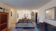 This single-floor residence is designed around an open-concept layout that connects the living and dining areas into one cohesive, functional space. The living room is carpeted throughout and accommodates generous seating, anchored by a central sofa arrangement. A ceiling fan with integrated lighting is centrally positioned overhead, providing both airflow and ambient illumination across the shared space.

The dining area sits directly behind the living room, furnished with a round table and chairs beneath framed wall art, contributing a casual yet defined eating space. A wooden display cabinet along one wall adds both storage and warmth to the interior aesthetic.

Neutral white walls and light-toned carpet run consistently throughout, creating a clean, understated backdrop. A hallway visible from the living area leads toward additional rooms, with a bright doorway suggesting natural light deeper within the unit. Nearby, a built-in closet with open shelving and a dark entry door marks a dedicated storage or entryway zone.

Overall, the space presents a practical and approachable layout with a calm, neutral ambiance suited for everyday living.