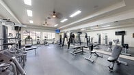 Located in San Antonio, Texas, this fitness center offers a comprehensive exercise facility designed to support a wide range of workout routines. The space is laid out on a single floor and provides ample room for both cardio and strength training activities.

The exercise room features durable rubber flooring throughout, providing a safe and functional surface for all types of workouts. A full wall of mirrors lines one side of the room, enhancing the sense of space and allowing users to monitor their form during exercise. Recessed fluorescent lighting and dual ceiling fans ensure the space remains bright and well-ventilated.

The cardio section is equipped with multiple treadmills and stair climbers, while the strength training area includes a variety of cable machines, weight benches, and plate-loaded equipment from brands such as Matrix. A dedicated free weights station features a tiered dumbbell rack with a full range of weights, accompanied by an adjustable bench.

Wall-mounted flat-screen televisions are positioned on opposing walls, providing entertainment during workouts. Large windows along one wall allow natural light to fill the room, and glass-paneled doors connect the fitness center to an adjacent outdoor area. A fire extinguisher and clearly marked exit signage reflect attention to safety standards throughout the facility.