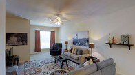 This single-floor space showcases a thoughtfully arranged living room that serves as its central feature. The room is finished with smooth, light-colored walls and a neutral ceiling equipped with a ceiling fan with integrated lighting, providing both air circulation and ambient illumination. A window dressed with warm rust-colored curtains allows natural light to filter into the space, contributing to its inviting atmosphere.

The living area is furnished with a plush sofa accented by an assortment of decorative throw pillows, complemented by a matching sectional and a classic dark leather recliner chair. A rectangular coffee table anchors the seating arrangement, resting atop a large, ornate area rug with circular medallion patterns in neutral and earth tones that adds visual interest to the floor.

Ambient lighting is provided by two table lamps with warm-toned shades, positioned to create a balanced, welcoming glow throughout the room. A large canvas print depicting the Golden Gate Bridge serves as a prominent wall feature, adding a bold decorative focal point. The overall ambiance is comfortable and casual, reflecting a well-organized and livable interior.