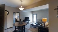 This single-floor space offers a thoughtfully designed open-concept layout that seamlessly connects the dining and living areas, creating a cohesive and functional environment. The dining area is anchored by a rich, dark-stained round table paired with elegant wrought iron accents, illuminated overhead by a classic pendant light fixture with a frosted glass shade. The living area flows naturally from the dining space, featuring a plush area rug, a comfortable sofa with decorative upholstery, and a sleek media console housing a flat-screen television. A large door with glass panels allows natural light to fill the space, brightening the neutral gray walls and light-toned flooring. Decorative table lamps add warm, ambient lighting throughout. A coffered ceiling detail adds subtle architectural interest and dimension to the shared living and dining zone. Adjacent to the main living area, a glimpse into the bedroom reveals a cozy, well-furnished retreat with a full-size bed and coordinating décor. The overall ambiance is warm and inviting, with a consistent dark wood and neutral color palette that lends the space a cohesive, modern yet comfortable aesthetic throughout.