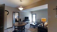 This single-floor space offers a thoughtfully designed open-concept layout that seamlessly connects the dining and living areas, creating a cohesive and functional environment. The dining area is anchored by a rich, dark-stained round table paired with elegant wrought iron accents, illuminated overhead by a classic pendant light fixture with a frosted glass shade. The living area flows naturally from the dining space, featuring a plush area rug, a comfortable sofa with decorative upholstery, and a sleek media console housing a flat-screen television. A large door with glass panels allows natural light to fill the space, brightening the neutral gray walls and light-toned flooring. Decorative table lamps add warm, ambient lighting throughout. A coffered ceiling detail adds subtle architectural interest and dimension to the shared living and dining zone. Adjacent to the main living area, a glimpse into the bedroom reveals a cozy, well-furnished retreat with a full-size bed and coordinating décor. The overall ambiance is warm and inviting, with a consistent dark wood and neutral color palette that lends the space a cohesive, modern yet comfortable aesthetic throughout.