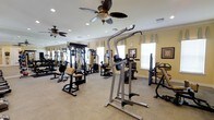This fitness center offers a comprehensive workout environment designed to accommodate a variety of exercise routines. The space is outfitted with an extensive selection of strength-training equipment, including cable machines, seated press stations, a power tower for bodyweight exercises, and various resistance machines arranged throughout the room. The layout provides ample space between equipment, allowing for comfortable movement and safe use of all stations. Neutral-toned carpet covers the floor, and the warm yellow walls contribute to an inviting atmosphere. Multiple ceiling fans with decorative wooden blades are installed overhead, complementing the recessed lighting to ensure the space remains well-lit and properly ventilated. Natural light filters in through several windows dressed with tailored valances, adding brightness to the environment. Wall-mounted mirrors are positioned along one side, a practical feature that allows users to monitor their form during workouts. Framed artwork on the walls adds a polished, community-oriented feel to the space. Overall, the fitness center strikes a balance between functional design and comfortable aesthetics, making it a welcoming facility for a range of fitness levels and workout goals.