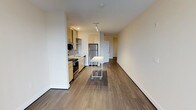 This single-floor space showcases a clean, contemporary design with an open-concept layout that seamlessly connects the kitchen and living areas. The kitchen is well-appointed with stainless steel appliances, including a refrigerator, range, and microwave, complemented by light wood cabinetry that adds a warm, modern aesthetic. A mobile kitchen island provides flexible workspace and additional functionality. Recessed and track lighting illuminate the kitchen area, while the open living space benefits from smooth white walls and neutral tones that create a bright, airy ambiance. Warm hardwood-style flooring runs continuously throughout the unit, lending a cohesive and inviting feel underfoot. The generous layout offers an open sightline from the kitchen through to the living area and toward what appears to be a hallway leading to additional rooms, suggesting a practical and well-organized floor plan. The combination of contemporary finishes, functional design, and neutral color palette makes this a versatile space well-suited for a variety of uses and furnishing styles.