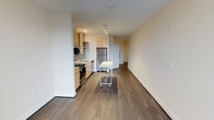 This single-floor space showcases a clean, contemporary design with an open-concept layout that seamlessly connects the kitchen and living areas. The kitchen is well-appointed with stainless steel appliances, including a refrigerator, range, and microwave, complemented by light wood cabinetry that adds a warm, modern aesthetic. A mobile kitchen island provides flexible workspace and additional functionality. Recessed and track lighting illuminate the kitchen area, while the open living space benefits from smooth white walls and neutral tones that create a bright, airy ambiance. Warm hardwood-style flooring runs continuously throughout the unit, lending a cohesive and inviting feel underfoot. The generous layout offers an open sightline from the kitchen through to the living area and toward what appears to be a hallway leading to additional rooms, suggesting a practical and well-organized floor plan. The combination of contemporary finishes, functional design, and neutral color palette makes this a versatile space well-suited for a variety of uses and furnishing styles.