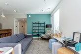 This contemporary single-floor space is thoughtfully designed with a clean, modern aesthetic and a functional layout. The kitchen area is a standout feature, showcasing white cabinetry with under-shelf LED lighting, dark countertops, and a full suite of stainless steel appliances including a full-size refrigerator and microwave. A warm wood-paneled island with bar seating beneath pendant lighting serves as a central gathering point, anchoring the kitchen to the open flow of the space. Polished concrete floors run throughout, lending an industrial-chic character that ties the areas together seamlessly. The lounge area offers a welcoming retreat, anchored by a bold teal accent wall that adds a pop of color against the neutral grey surroundings. A comfortable grey sofa, an armchair, and a variety of seating options are arranged around a marble-topped coffee table on a textured area rug. Open black metal shelving displays curated décor, while a work desk and colorful ottomans provide a versatile, creative corner. Large windows fitted with blinds allow generous natural light to filter in, offering views of the surrounding greenery and providing an airy, bright ambiance throughout the space.