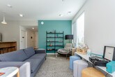 This contemporary single-floor space is thoughtfully designed with a clean, modern aesthetic and a functional layout. The kitchen area is a standout feature, showcasing white cabinetry with under-shelf LED lighting, dark countertops, and a full suite of stainless steel appliances including a full-size refrigerator and microwave. A warm wood-paneled island with bar seating beneath pendant lighting serves as a central gathering point, anchoring the kitchen to the open flow of the space. Polished concrete floors run throughout, lending an industrial-chic character that ties the areas together seamlessly. The lounge area offers a welcoming retreat, anchored by a bold teal accent wall that adds a pop of color against the neutral grey surroundings. A comfortable grey sofa, an armchair, and a variety of seating options are arranged around a marble-topped coffee table on a textured area rug. Open black metal shelving displays curated décor, while a work desk and colorful ottomans provide a versatile, creative corner. Large windows fitted with blinds allow generous natural light to filter in, offering views of the surrounding greenery and providing an airy, bright ambiance throughout the space.